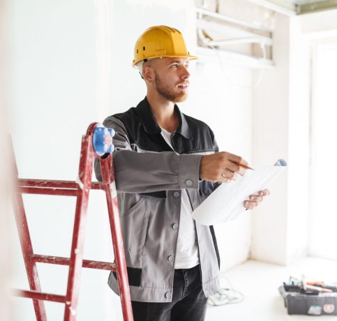 Young engineer in work clothes and yellow hardhat dreamily looking aside holding plan of new apartments in hands with red ladder near in repairing flat